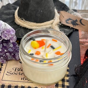 May include: A white candle in a glass jar with a black witch's hat, a purple amethyst crystal, and a black and white checkered fabric. The candle is decorated with candy corn, sprinkles, and a wooden wick.
