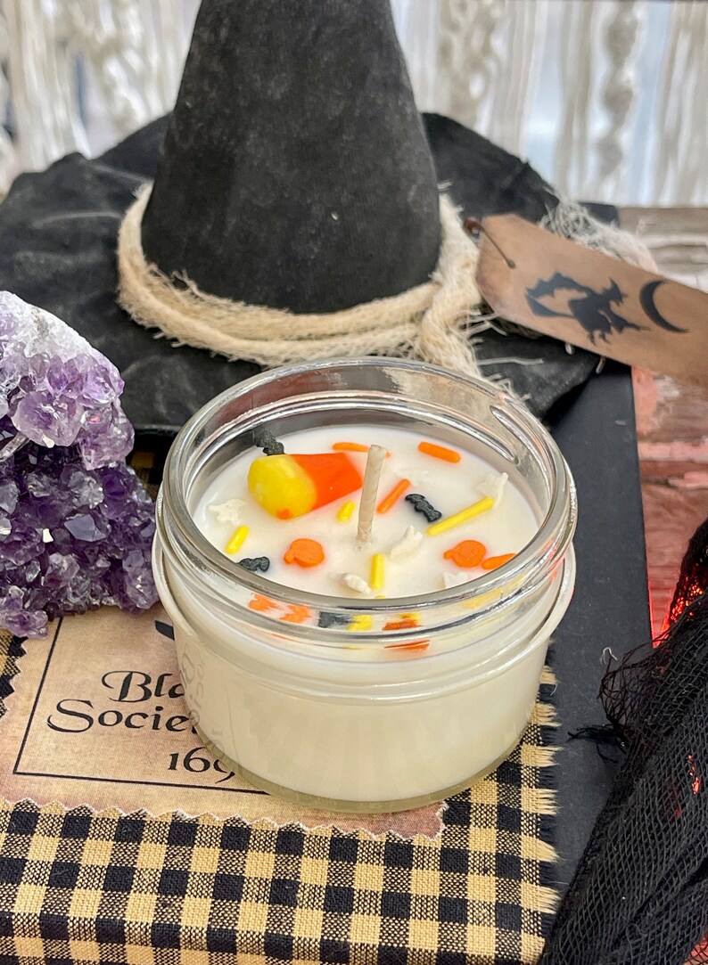 May include: A white candle in a glass jar with a black witch's hat, a purple amethyst crystal, and a black and white checkered fabric. The candle is decorated with candy corn, sprinkles, and a wooden wick.