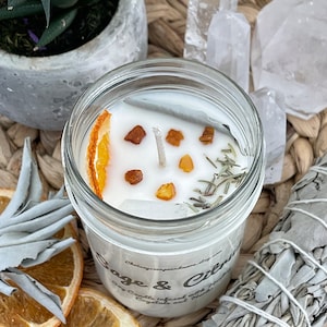 May include: A white soy candle in a glass jar with a dried orange slice, herbs, and crystals. The candle is labeled "Sage & Citrus" and "Soy Candle Infused with Gemstones and Herbs".