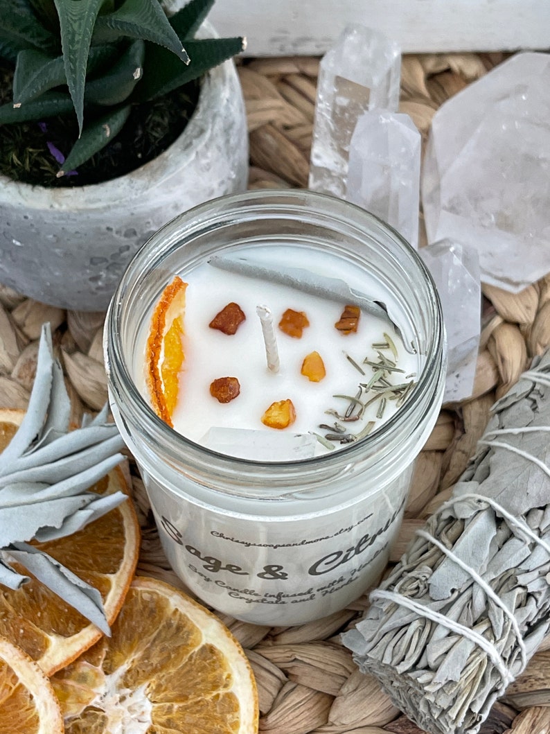 May include: A white soy candle in a glass jar with a dried orange slice, herbs, and crystals. The candle is labeled "Sage & Citrus" and "Soy Candle Infused with Gemstones and Herbs".