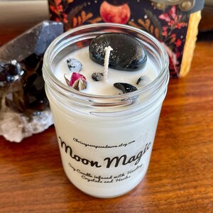 May include: A white soy candle in a glass jar with a black label that reads "Moon Magic Soy Candle Infused with Healing Crystals and Herbs" and is decorated with black crystals and a dried rose.