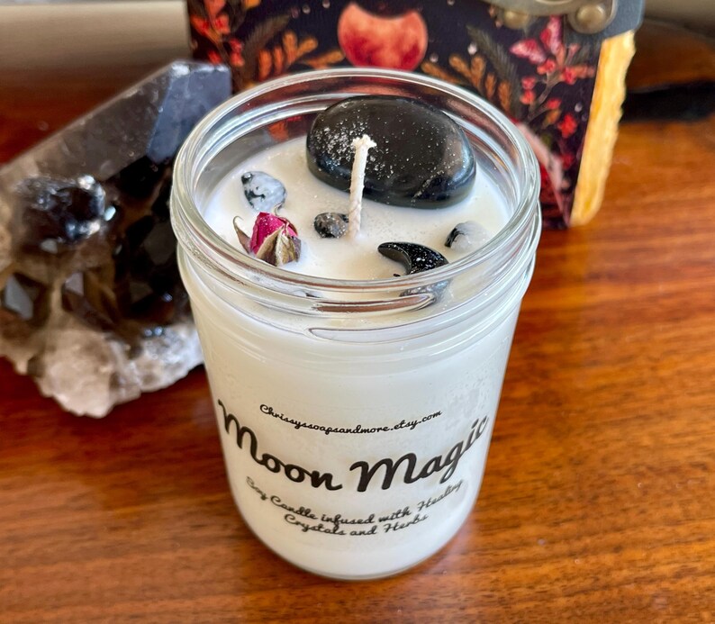May include: A white soy candle in a glass jar with a black label that reads "Moon Magic Soy Candle Infused with Healing Crystals and Herbs" and is decorated with black crystals and a dried rose.