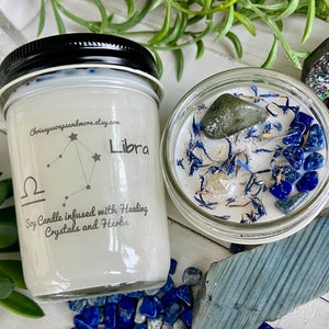 May include: A white soy candle with a black lid and a label that says "Libra" and "Soy Candle infused with Healing Crystals and Herbs". The candle is surrounded by blue and green crystals.