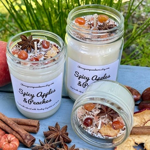 May include: Three glass jars filled with soy candles. The candles are topped with dried fruit, cinnamon sticks, and star anise. The labels on the jars read "Spicy Apples & Peaches".