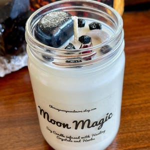 May include: A white soy candle in a glass jar with a black label that reads "Moon Magic Soy Candle infused with Healing Crystals and Herbs". The candle is topped with black and white crystals.