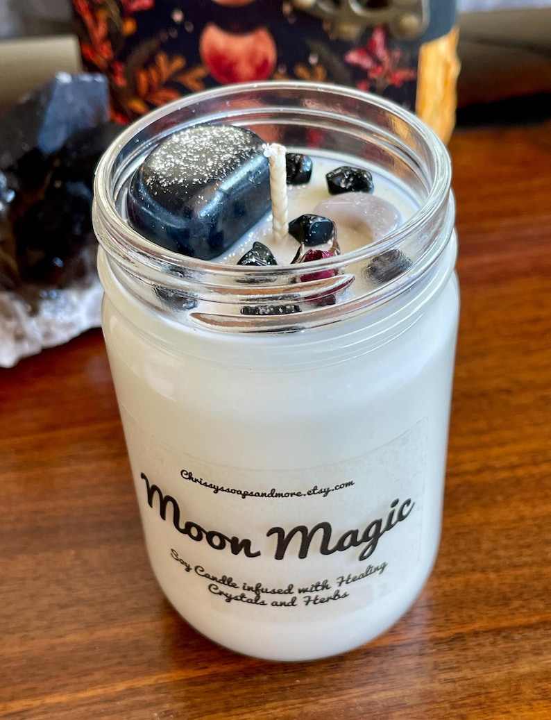 May include: A white soy candle in a glass jar with a black label that reads "Moon Magic Soy Candle infused with Healing Crystals and Herbs". The candle is topped with black and white crystals.