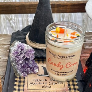 May include: A glass jar candle with a white soy wax base and a label that reads "Chrissy's Candle Creations Hand Poured in Dillsburg PA Candy Corn Chrissyssoapsandmore.etsy.com Soy Candle". The candle is decorated with orange, yellow, and white candy corn pieces and black sprinkles. The candle is sitting on a black and white checkered surface with a purple amethyst crystal and a black witch's hat.