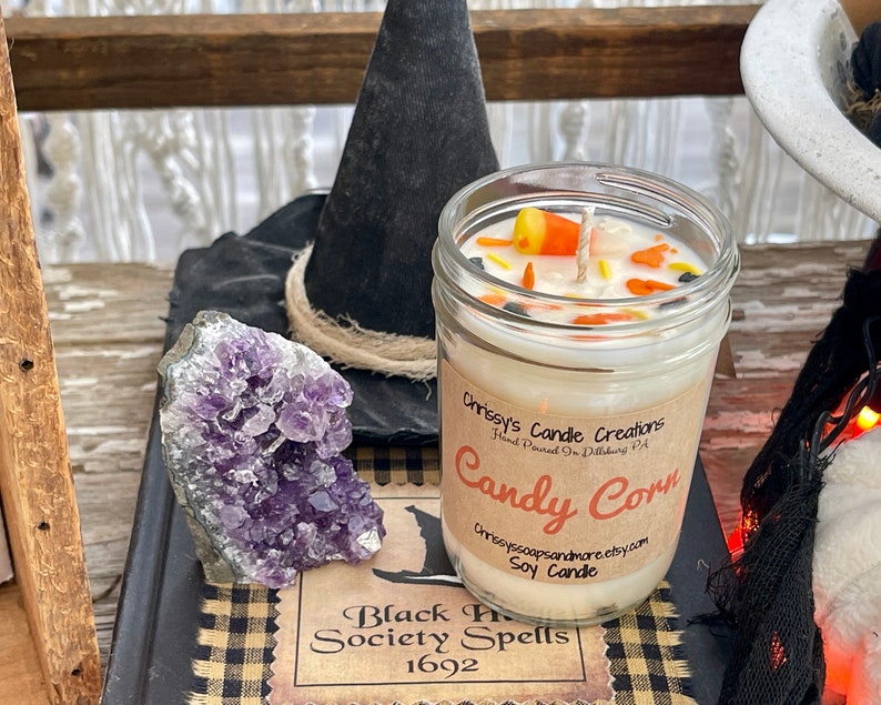 May include: A glass jar candle with a white soy wax base and a label that reads "Chrissy's Candle Creations Hand Poured in Dillsburg PA Candy Corn Chrissyssoapsandmore.etsy.com Soy Candle". The candle is decorated with orange, yellow, and white candy corn pieces and black sprinkles. The candle is sitting on a black and white checkered surface with a purple amethyst crystal and a black witch's hat.