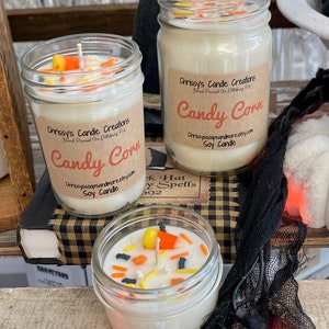 May include: Three glass jars with white soy candles decorated with candy corn and Halloween sprinkles. The jars have labels that say "Chrissy's Candle Creations" and "Candy Corn".