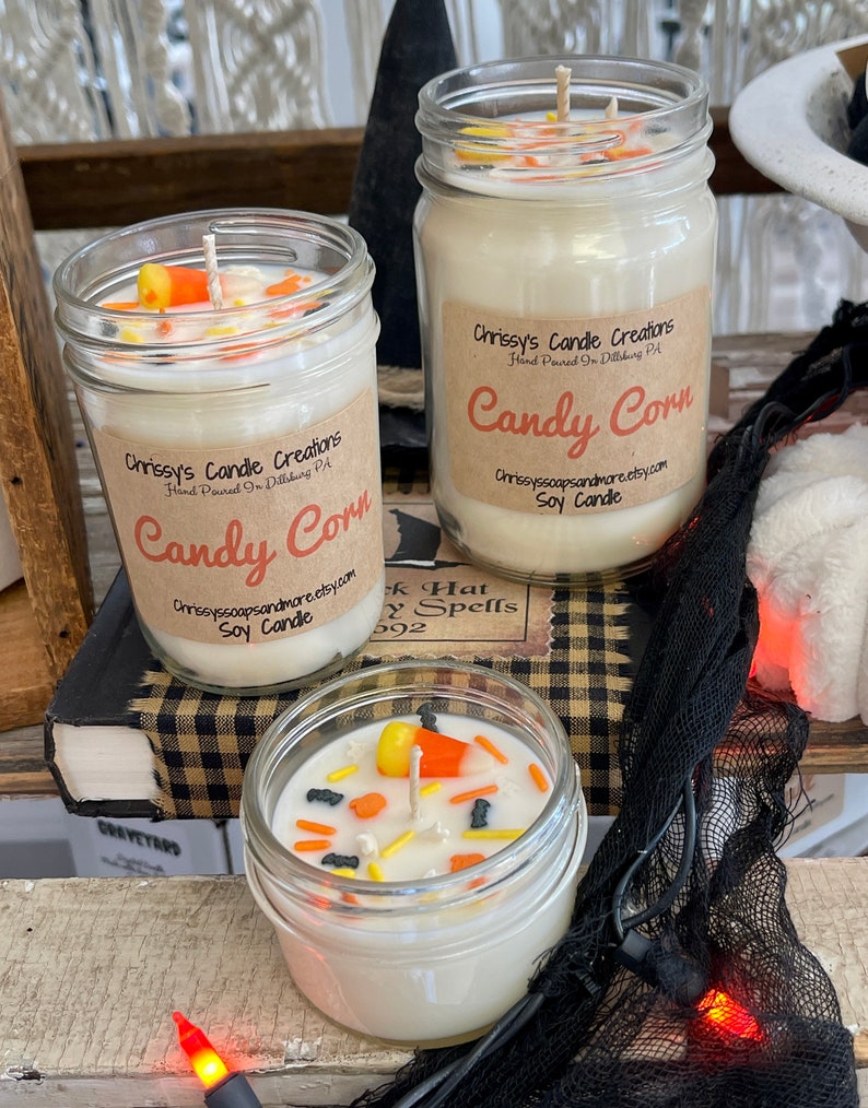 May include: Three glass jars with white soy candles decorated with candy corn and Halloween sprinkles. The jars have labels that say "Chrissy's Candle Creations" and "Candy Corn".