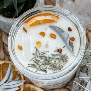 May include: A white candle with dried orange peel, sage, and herbs in a clear glass jar. The candle is surrounded by white crystals and dried sage.