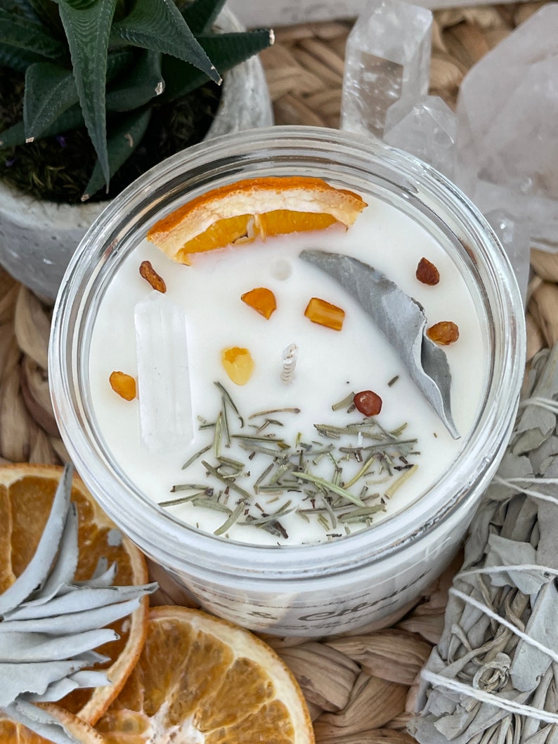 May include: A white candle with dried orange peel, sage, and herbs in a clear glass jar. The candle is surrounded by white crystals and dried sage.