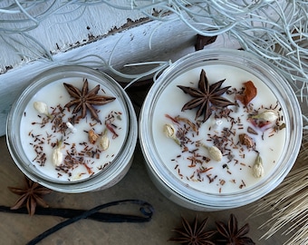 May include: Two glass jars filled with white soy wax candles. The candles are topped with star anise, dried flowers, and other botanicals.