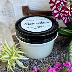 May include: A white jar with a black lid labeled "Relaxation with Essential Oils, Crystals and Herbs". The jar is surrounded by dried lavender flowers and green plants.
