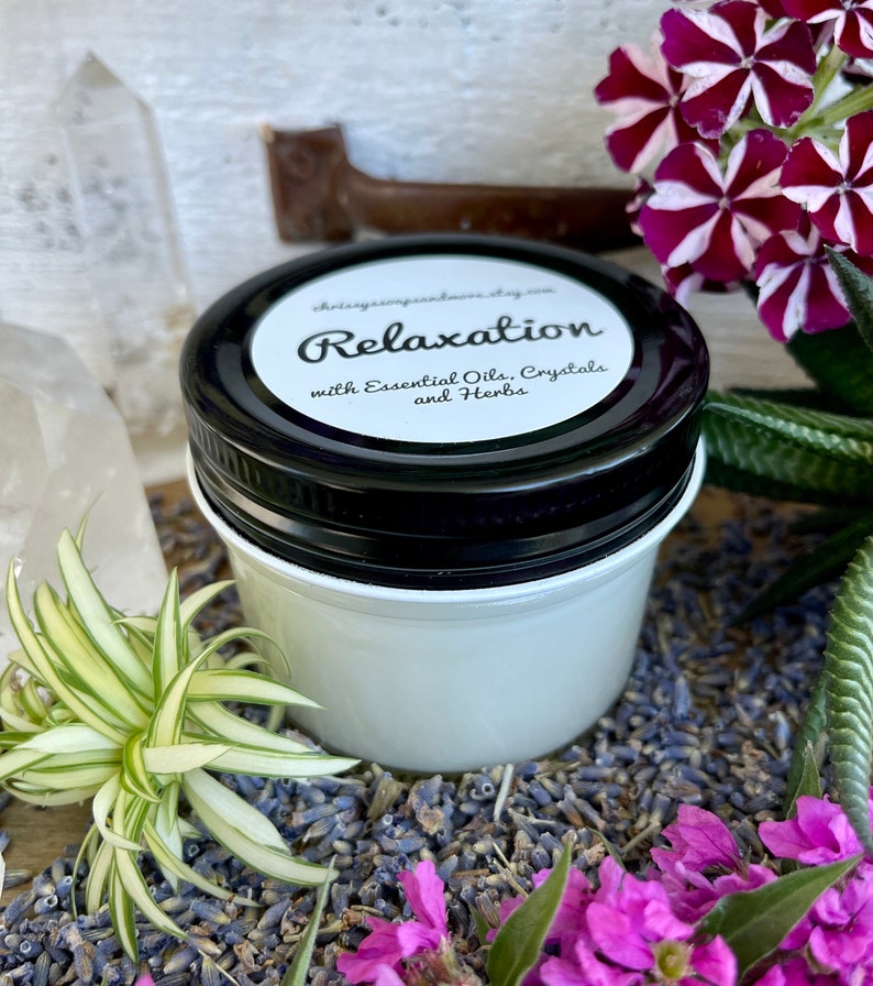 May include: A white jar with a black lid labeled "Relaxation with Essential Oils, Crystals and Herbs". The jar is surrounded by dried lavender flowers and green plants.