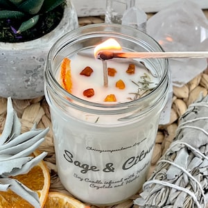 May include: A clear glass jar candle with a white soy wax base. The candle is infused with dried orange slices, herbs, and crystals. The label reads "Sage & Citrus Soy Candle infused with Herbs, Crystals and Flowers".