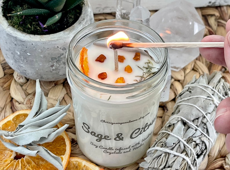 May include: A clear glass jar candle with a white soy wax base. The candle is infused with dried orange slices, herbs, and crystals. The label reads "Sage & Citrus Soy Candle infused with Herbs, Crystals and Flowers".