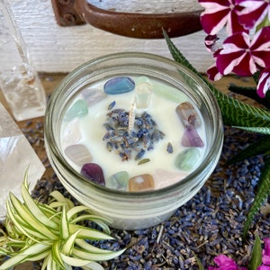 May include: A white candle with lavender and colorful crystals in a clear glass jar. The candle is surrounded by dried lavender flowers.