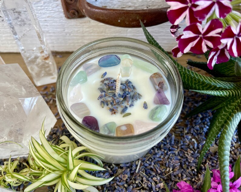 May include: A white candle with lavender and colorful crystals in a clear glass jar. The candle is surrounded by dried lavender flowers.
