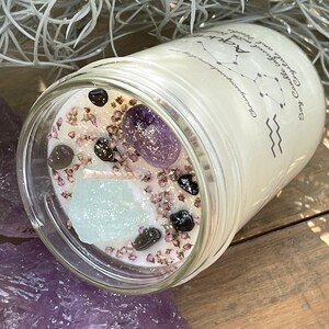 Peut inclure: Une bougie blanche dans un pot en verre avec un couvercle transparent. La bougie pr&eacute;sente un motif de constellation et le texte "Soy Candle with Crystals and Herbs for Aquarius". Le pot contient de petits cristaux et des fleurs s&eacute;ch&eacute;es.
