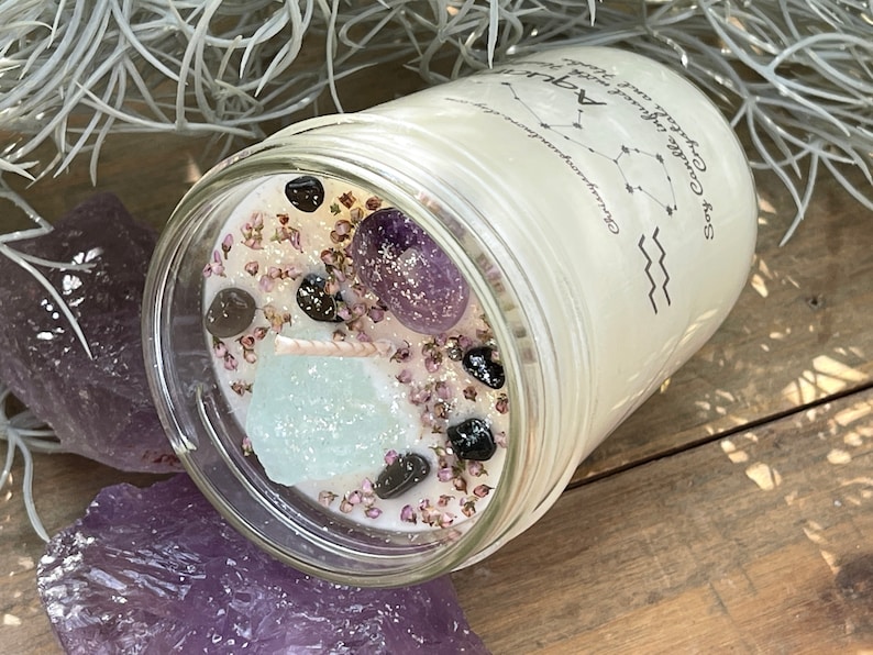 Peut inclure: Une bougie blanche dans un pot en verre avec un couvercle transparent. La bougie pr&eacute;sente un motif de constellation et le texte "Soy Candle with Crystals and Herbs for Aquarius". Le pot contient de petits cristaux et des fleurs s&eacute;ch&eacute;es.