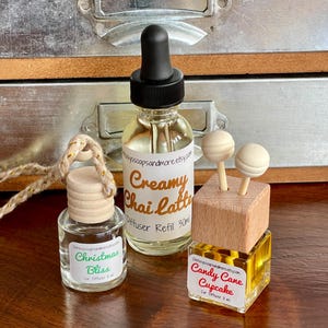 May include: Three car air fresheners with different scents. One is a clear bottle with a black dropper and the label says "Creamy Chai Latte". Another says "Christmas Bliss". The third says "Candy Cane Cupcake".