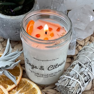 May include: A white soy candle with a sage and citrus scent. The candle is in a clear glass jar with a white label that says "Sage & Citrus Soy Candle infused with Healing Crystals and Herbs". The candle is surrounded by dried orange slices and a bundle of white sage.