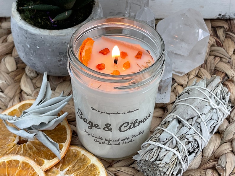 May include: A white soy candle with a sage and citrus scent. The candle is in a clear glass jar with a white label that says "Sage & Citrus Soy Candle infused with Healing Crystals and Herbs". The candle is surrounded by dried orange slices and a bundle of white sage.