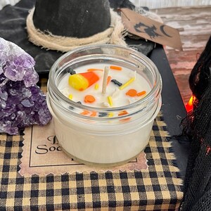 May include: A white candle in a clear glass jar with a black and orange candy corn, black bats, and orange sprinkles on top. The candle is sitting on a black and white checkered surface.