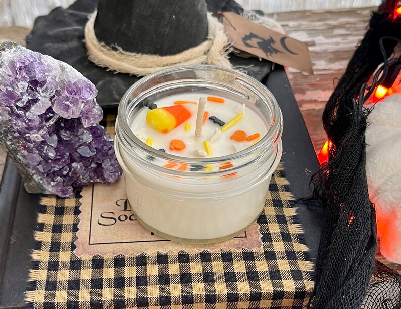 May include: A white candle in a clear glass jar with a black and orange candy corn, black bats, and orange sprinkles on top. The candle is sitting on a black and white checkered surface.