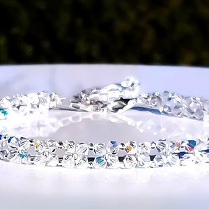 May include: A delicate silver bracelet with clear, faceted crystals. The crystals are arranged in a repeating pattern, reflecting light and creating a sparkling effect. The bracelet is on a white surface.
