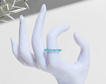 May include: A ring featuring a row of small, round, light blue faceted stones set in a thin, metallic band. The stones exhibit a bright, reflective quality, creating a consistent sparkle across the entire circumference of the circular jewelry piece.