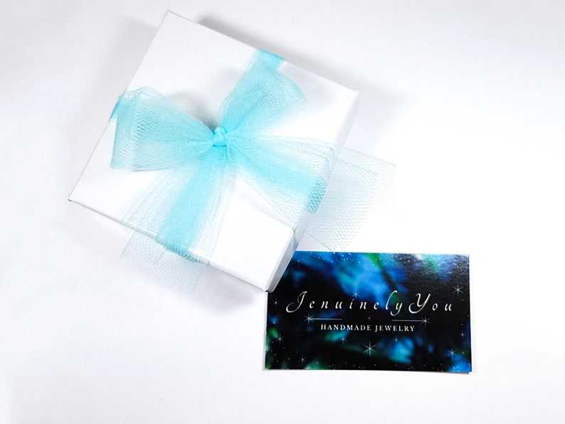 A small square white gift box with turquoise mesh ribbon wrapped into a bow sits next to a business card with a dark multicolored background with the words "JenuinelyYou Handmade Jewelry" written on it in white.