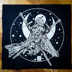 May include: Black and white embroidered patch of a bird with two swords crossed behind it, set against a crescent moon and starry sky. The patch is on a black fabric background.