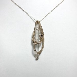 Puede incluir: Un colgante de cristal de cuarzo transparente envuelto en alambre de color dorado, suspendido de una delicada cadena de plata. El trabajo de alambre presenta intrincados remolinos y una pequeña gema de color rojo oscuro. El cristal mide unos 7.6 cm de largo.