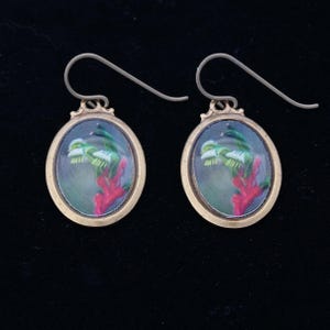 May include: A pair of gold-toned earrings with a green and red floral design inside a clear oval frame.
