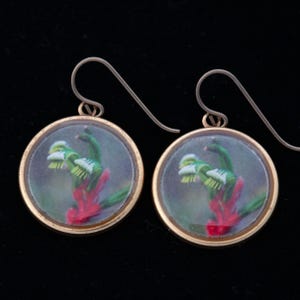 May include: A pair of gold-toned earrings with a round, clear glass pendant featuring a red and green flower design.