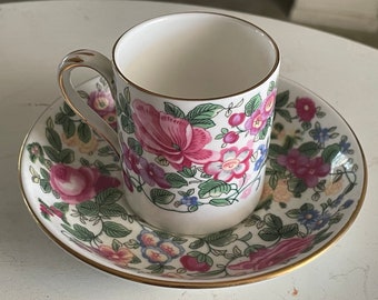 May include: A white teacup and saucer with a floral pattern in shades of pink, green, and yellow. The cup has a gold rim and the saucer has a gold band around the edge.