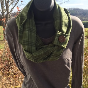 May include: A green and white plaid infinity scarf with a wooden button. The scarf is worn on a mannequin with a gray long-sleeved shirt.