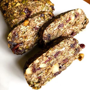 May include: A loaf of gluten-free seed bread sliced into three pieces, featuring a dense texture with visible green pumpkin seeds, sunflower seeds, and dark red dried cranberries. The text "Gluten-Free Ultimate Seed Bread" is printed in red.
