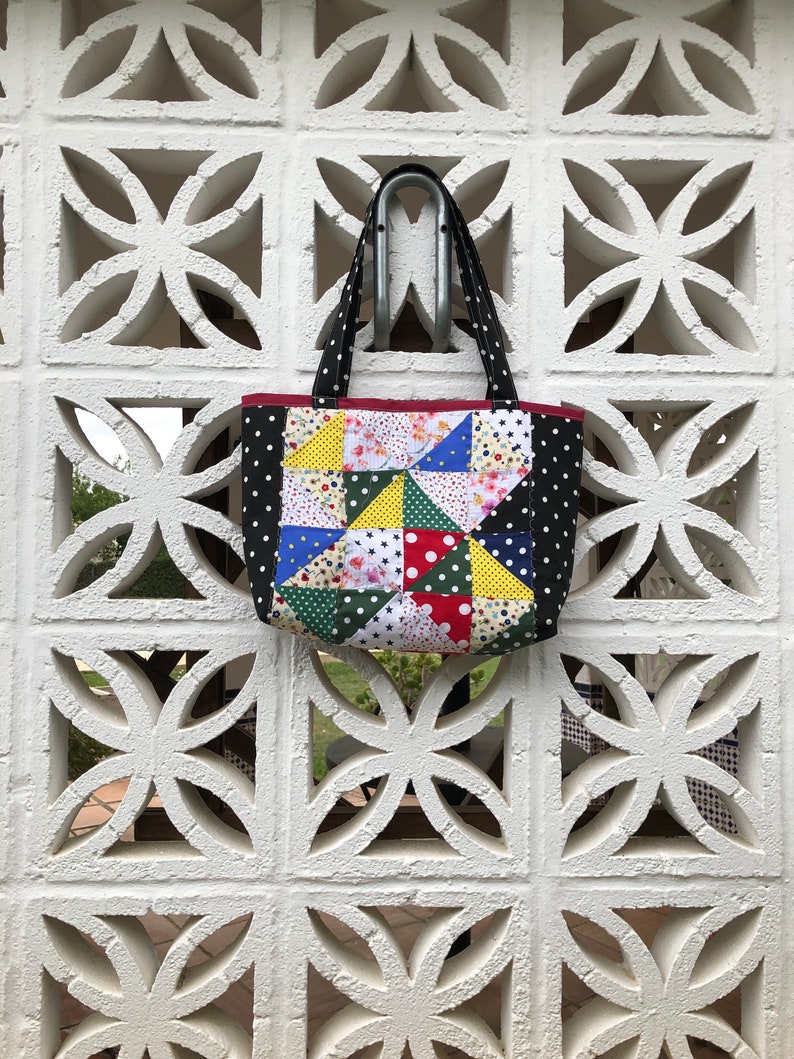 Pu&ograve; includere: Una borsa a mano a pois neri e bianchi con un design a patchwork con vari colori e motivi, tra cui rosso, giallo, verde, blu e rosa. La borsa ha un'unica maniglia ed &egrave; appesa a un muro bianco.