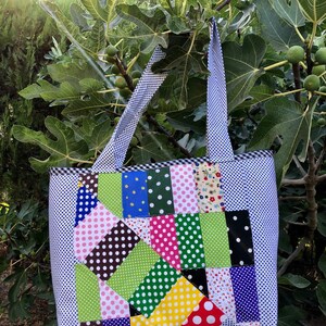 Pu&ograve; includere: Una borsa tote in patchwork con sfondo bianco e quadrati di tessuto colorati con pois, strisce e motivi floreali. La borsa ha due manici e un bordo a quadri bianco e blu.