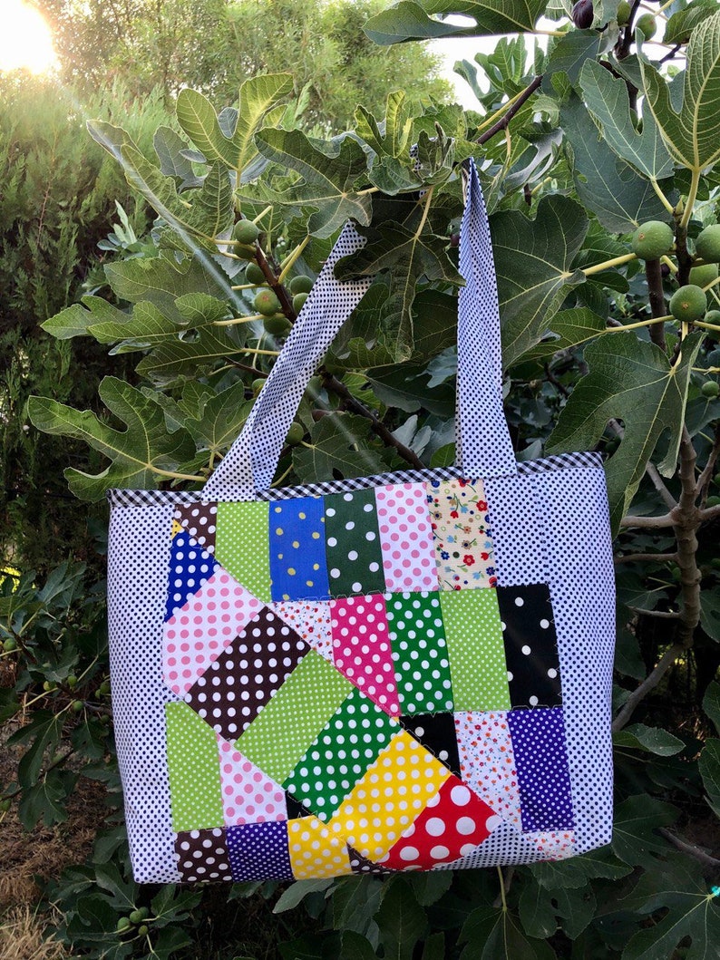 Pu&ograve; includere: Una borsa tote in patchwork con sfondo bianco e quadrati di tessuto colorati con pois, strisce e motivi floreali. La borsa ha due manici e un bordo a quadri bianco e blu.