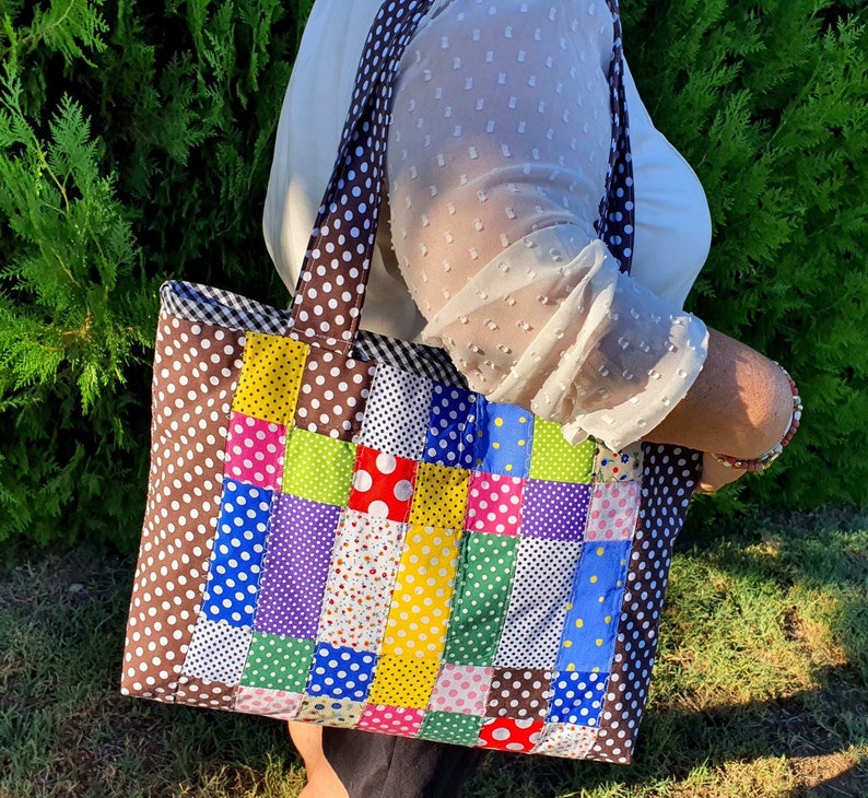 Pu&ograve; includere: Una borsa a mano in patchwork marrone e bianco con quadrati e pois colorati. La borsa ha un manico in tessuto marrone e una chiusura a zip.