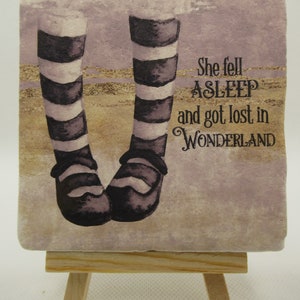 May include: A decorative tile featuring a whimsical illustration of striped legs and black shoes. The tile has a light purple background with gold glitter accents. The text on the tile reads "She fell asleep and got lost in WONDERLAND".