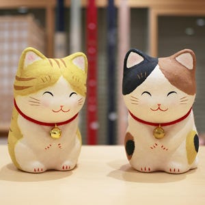 以下が含まれることがあります： 2つのセラミック製招き猫の置物。1つは黄色の茶色の縞模様、もう1つは白と黒と茶色のパッチがあります。どちらも赤い首輪に金の鈴と笑顔の顔があります。明るい茶色の表面に座っています。
