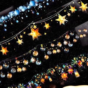 May include: A black background with multiple strings of lights. The lights are a mix of gold star shapes and round, clear bulbs. The lights are arranged in a cascading pattern, with the top row being the most distant and the bottom row being the closest. The lights are all lit and glowing.