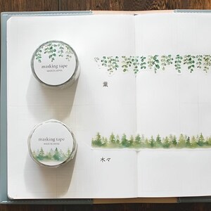 Può includere: Due rotoli di nastro washi con disegni ad acquerello verdi. Il rotolo superiore presenta un motivo a ghirlanda di foglie con il testo "masking tape made in Japan". Il rotolo inferiore presenta una scena di bosco con il testo "masking tape made in Japan".