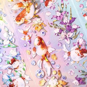 K&ouml;nnte beinhalten: Eine Bogen mit bunten Aufklebern mit Goldfischen und Blumen. Die Aufkleber befinden sich auf einem wei&szlig;en Hintergrund mit einem Regenbogeneffekt. Die Goldfische sind orange und wei&szlig; mit schwarzen Augen. Die Blumen sind lila, rosa und wei&szlig;.