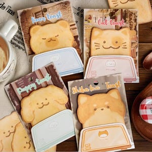 May include: A collection of cute, illustrated toast-shaped memo pads featuring animal faces: a bear, cat, puppy, and Shiba Inu. Each pad has a matching toaster-shaped holder. The text "Bear toast", "Cat toast", "Puppy toast", and "Shiba Inu Toast" is visible.